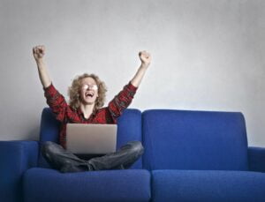 Photo of Excited Person With Hands Up Sitting on A Blue Sofa While Using a Laptop After Winning a Daily Entry Sweepstakes